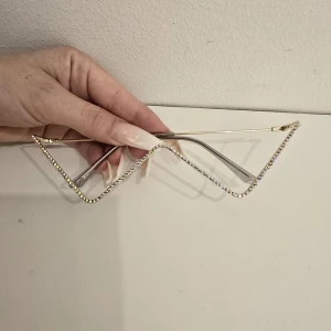 Glittriga kantiga glasögon i guld - Unika glasögon med kantig, zickzack-formad ram i guldmetall och glittrande stenar längs hela bågen. Perfekt statement piece för dig som vill sticka ut och glänsa. Ramen är tunn och lätt, utan glas i bågen.