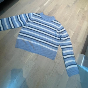 Randig blå stickad tröja - Säljer en stickad tröja med breda ränder i olika blå nyanser och vitt. Tröjan har ribbade muddar vid ärmslut, nederkant och halsringning.