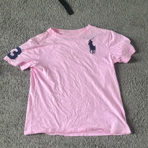 Rosa t-shirt från Ralph Lauren - Snygg rosa t-shirt från Ralph Lauren med stort mörkblått logotryck på bröstet och en blå siffra 3 på ärmen. Klassisk passform och rund halsringning. Perfekt för dig som gillar stilrena plagg med sportig touch.