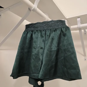 Mörkgröna satinskjorts med spets - Mörkgröna shorts i glansigt satinmaterial med bred spetskant upptill. Luftig och lös passform, perfekta för en chic och bekväm look. Snyggt fall och elegant känsla.