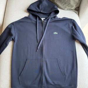 Marinblå hoodie från Lacoste  - Snygg marinblå hoodie från Lacoste i 100% bomull. Klassisk design med dragkedja, huva med snörning och känguruficka framtill. Ikoniska gröna krokodilloggan broderad på bröstet. Perfekt för en chill och stilren look.