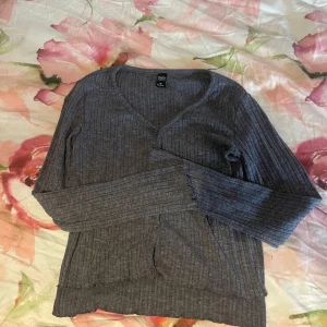 Grå ribbad kofta från Dazy - Mysig grå kofta från Dazy i ribbat material med v-ringning och knappar framtill. Långärmad och i en normal passform, perfekt att slänga över en topp. Enkel och stilren design som funkar till det mesta.