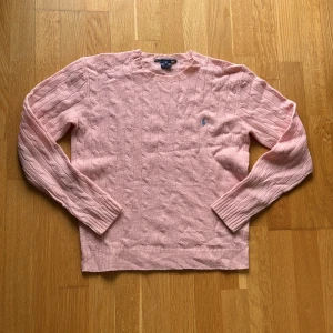 Rosa kabelstickad tröja från Ralph Lauren - Säljer en ljusrosa kabelstickad tröja från Ralph Lauren med klassisk rund halsringning och broderad logga på bröstet. Tröjan har långa ärmar och ribbade muddar, perfekt för dig som gillar en preppy och stilren look.