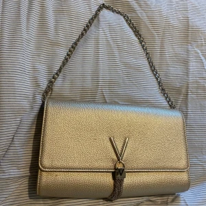 Guldig axelväska från Valentino - Säljer en snygg guldig axelväska från Valentino med kedjerem och ikoniskt V-spänne framtill med hängande kedjedetalj. Väskan har struktur i materialet, magnetlås och innerfack med dragkedja. Perfekt för dig som vill sticka ut med en lyxig accessoar. Måttet är 27/16cm.