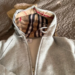 Grå hoodie från Burberry med dragkedja - Snygg grå hoodie från Burberry med klassiskt rutigt foder i huvan. Tröjan har dragkedja framtill, kängurufickor och snörning i huvan. Perfekt för dig som gillar stilrena och exklusiva plagg med en twist.