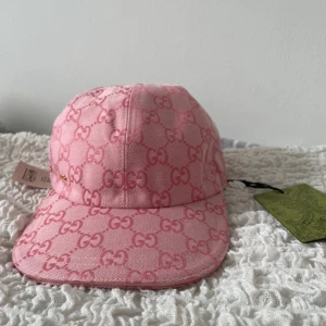 Rosa keps från Gucci med GG-mönster - Snygg rosa keps från Gucci med ikoniskt GG-mönster över hela kepsen. Tillverkad i bomullstyg och har en klassisk böjd skärm. Kepsen har en liten gulddetalj på sidan och originaletiketter kvar. Perfekt för dig som vill sticka ut med en lyxig accessoar.