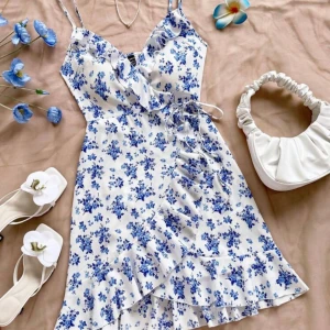 Vit och blå blommig omlottklänning - Superfin kort klänning med blått blommönster på vit botten. Klänningen har smala axelband, omlott-detalj med knyt i midjan och volang längs nederkanten. Perfekt för sommarens alla tillfällen och ger en fräsch och romantisk vibe. Aldrig använd!❤️