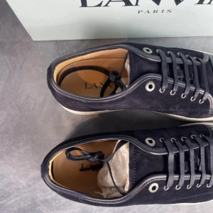 Svarta sneakers från Lanvin i mocka - Stilrena svarta sneakers från Lanvin med ovandel i mocka och tåparti i skinn. Skorna har vita platta sulor, rund tå och klassisk snörning. Perfekta för dig som gillar lyxiga och minimalistiska detaljer.