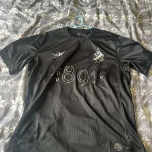Svart AIK fotbollströja Nike L - AIK fotbollströja i svart från Nike med Dri-FIT-material. Tryck med '1891' på bröstet, AIK:s klubbmärke och Nike-logga i vitt. Rund halsringning och korta ärmar. Diskret mönster i tyget och texten 'SMOKINGLIRARE' på ryggen.