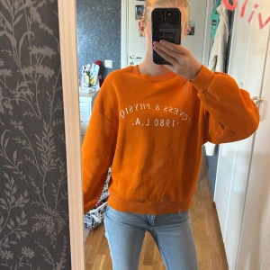 Orange sweatshirt från Gina Tricot XS - Superfräsch orange sweatshirt från Gina Tricot i storlek XS. Tröjan har trycket 'Fitness & Physique 1980 L.A.' framtill, rund hals och ribbade muddar vid ärmslut och nederkant. Perfekt för en chill och sportig look.