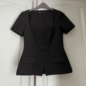 Svart blus med knappar från Zara - Stilren svart blus från Zara med korta ärmar och markerad midja. Djup, fyrkantig urringning och fem klädda knappar framtill. 