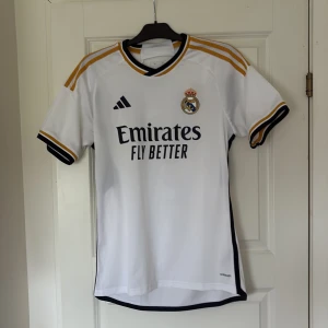 Real Madrid Bellingham 5 matchtröja - Säljer en officiell Real Madrid fotbollströja med Bellingham 5 på ryggen. Tröjan är vit med svarta och guldiga detaljer, Adidas-logga och klubbmärke på bröstet. Korta ärmar och tillverkad i lätt, ventilerande material. Perfekt för dig som älskar fotboll och Real Madrid. Inga fläckar eller något använt en gång säljer den för den är för stor för min son. Ny pris var 1300
