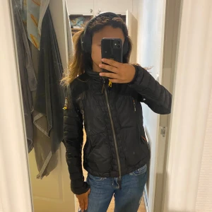 Svart Parajumpers pufferjacka - Säljer en svart pufferjacka från Parajumpers med orange detaljer och patch på ärmen. Jackan har hög krage, dragkedja framtill och flera fickor med dragkedja. Perfekt för kyliga dagar och riktigt snygg till jeans.