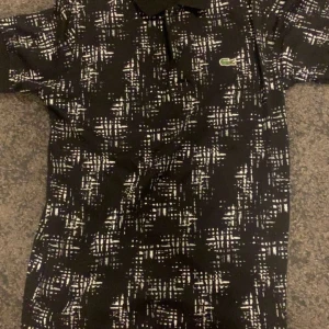 Svartvit pikétröja från Lacoste - Säljer en svart pikétröja från Lacoste med vitt abstrakt mönster. Klassisk krage med gröna och vita detaljer, knäppning framtill och den ikoniska gröna krokodilloggan på bröstet. Tröjan är i bomull och har korta ärmar. Perfekt för en stilren och sportig look.