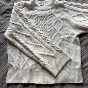 Vit kabelstickad tröja med mönster - Kabelstickad tröja i mjuk vit ullmix med grovt mönster och ribbade muddar. Klassisk rund halsringning och långärmad modell, perfekt för lager på lager under kyliga dagar. Snygg struktur som ger en stilren och mysig vibe.