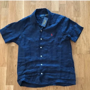 Mörkblå linneskjorta Polo Ralph Lauren - Snygg mörkblå kortärmad skjorta från Polo Ralph Lauren i klassisk passform. Skjortan är i svalt linne och har knappar framtill samt en bröstficka med röd broderad logga. Perfekt för varma dagar och stilrena outfits. Skriv gärna om ni har frågor 😄👍🏻
