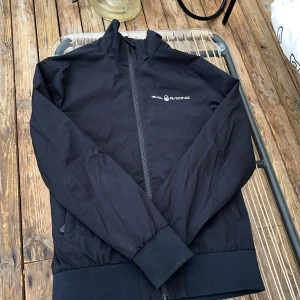 Svart vindjacka från Sail Racing - Svart vindjacka från Sail Racing med stilren design och logga på bröstet och ryggen. Jackan har hel dragkedja framtill, ribbade muddar och två sidofickor. Perfekt för dig som gillar sportig och clean look. Materialet är lätt och känns smidigt.