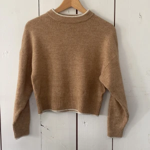 Beige stickad tröja från H&M - Supermysig beige stickad tröja från H&M i croppad modell. Tröjan har ribbade muddar och en rund halsringning med vit kant. Perfekt för lager-på-lager och chill dagar. Passar dig som gillar en enkel men snygg stil.