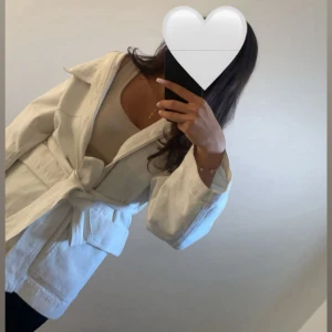 Vit jacka med bälte - Snygg krämvit jacka från Gina Tricot med bälte i midjan och stora fickor framtill. Jackan har klassisk krage, knappar och långa ärmar. Perfekt att slänga över en outfit för en clean och fräsch look. Har en liten defekt på ena armen (vet ej om det är jag som har gjort den eller om den fanns där när jag köpte den) men det är absolut inget som syns när man har på sig den!