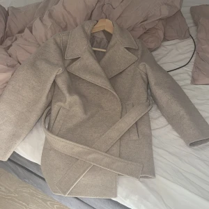 Beige kappa med bälte och krage - Stilren beige kappa med bred krage och bälte i midjan. Jackan har raka fickor och är helfodrad, perfekt för dig som vill ha en klassisk look med modern touch. Passar snyggt till både jeans och kostymbyxor.