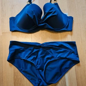 Mörkblå bikini med gulddetaljer XL - Snygg mörkblå bikini  i storlek XL. Överdelen har vadderade kupor och axelband med coola guldfärgade ringdetaljer, stl 95C. Bikinitrosan har hög midja och en twistad detalj framtill, stl XL. Materialet är mjukt och stretchigt syntet, perfekt för stranden. Eco.