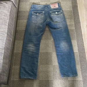 True Religion jeans - Säljer ett par blå jeans från True Religion med ikoniska kontrastsömmar och stora fickor med lock och knappar bak. Jeansen har coola slitningar framtill. Tillverkade i bomull och har detaljer som nitar och logopatch. Passformen är lite baggy men inte överdrivet. Hör av dig vad minsta fundering.