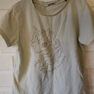 Ljusgrön t-shirt med enhörning H&M - Ljusgrön t-shirt från H&M med korta ärmar och ett stort tryck av en enhörning på framsidan samt texten 'Be strong and brave'. Skön och mjuk i bomull, perfekt för dig som gillar magiska motiv. Lätt tvättpåverkad 