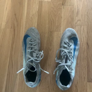 Nike grå-blå fotbollsskor med snörning elite  - Säljer ett par gråa Nike fotbollsskor med blå swoosh och detaljer. Skorna har snörning och är tillverkade i syntetmaterial med mönstrad ovandel. Sulan är vit med blå dobbar, perfekt för spel på gräs. Snygg och sportig design för dig som älskar fotboll.