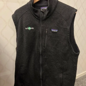 Svart fleeceväst från Patagonia - Svart fleeceväst från Patagonia med hel dragkedja framtill och hög krage. Västen har broderad logga på bröstet och en bröstficka med dragkedja. Perfekt för lager-på-lager och chill vardagsstil.