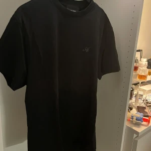 Svart t-shirt från Axel Arigato - Clean svart t-shirt från Axel Arigato. Klassisk passform med korta ärmar och diskret broderad logga på bröstet. Perfekt basic-plagg med snygg minimalistisk vibe. Aldrig använd