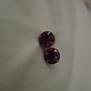 Guldiga örhängen med lila sten - Snygga örhängen i guldton med rund, gnistrande lila sten. Klassisk design med stift, perfekta för att ge lite extra färg till din look. Stenarna har en djup lila nyans som fångar ljuset superfint.