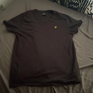 Svart t-shirt från Lyle & Scott - Svart t-shirt från Lyle & Scott med klassisk passform och korta ärmar. T-shirten har en gul broderad logga på bröstet och är tillverkad i mjuk bomull. Perfekt basic-plagg med stilren look.