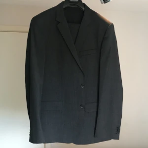 Mörkgrå kostym set från Hugo Boss - Snygg mörkgrå kostym från Hugo Boss med klassisk tvåknäppt kavaj och matchande byxor. Kavajen har smala slag, innerfickor och raka fickor med lock. Perfekt för dig som vill ha en stilren och tidlös look.
