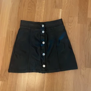 Svart kortkjol i skinnimitation H&M - Snygg svart kortkjol från H&M i skinnimitation med silvriga knappar framtill. Kjolen har en rak passform och markerad midja, perfekt till en cool och trendig look. Enkel att matcha med både stickade tröjor och t-shirts.