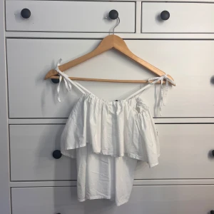 Vit volangig linnetopp med knytband - Supersöt vit linnetopp med volang och smala axelband som knyts på axlarna. Toppen har en luftig och flowy känsla, perfekt för varma dagar. Passar dig som gillar en romantisk och chill stil.