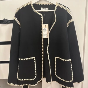 Svart cardigan jacka från AWARE XS - Svart cardigan jacka från AWARE med kontrasterande beige broderad kant runt hals, öppning, fickor och ärmslut. Jackan har två stora fickor framtill och en öppen design med bälte att knyta runt vid midjan . Helt ny med prislapp