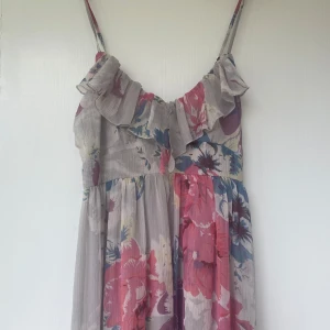 Lång blommig klänning med volang - Superfin långklänning med tunna axelband och volangdetalj upptill. Klänningen är ljusgrå med stora blommor i rosa, lila och blå toner. Tunt, flowy tyg som känns luftigt och perfekt för varma dagar. Sitter snyggt och ger en romantisk vibe.