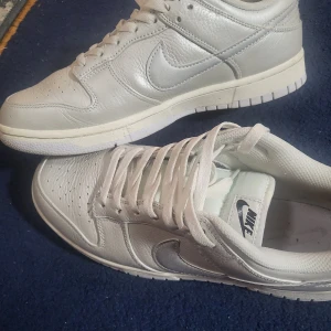 Nike Dunk Low i vitt och silver - Nike Dunk Low sneakers i vitt skinn med silverfärgad swoosh. Klassisk siluett med perforerad tå, vit platt sula och snörning. Skorna har en låg profil och är perfekta för dig som gillar stilrena och cleana sneakers med retrovibbar.