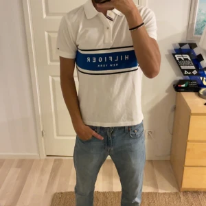 Vit Tommy Hilfiger pikétröja - Vit pikétröja från Tommy Hilfiger med blått och svart band över bröstet och tydlig logga. Klassisk krage med knappar och korta ärmar. Skön bomullskvalitet och stilren design som sticker ut med den blåa detaljen.