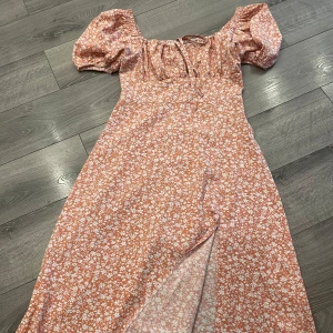 Blommig långklänning med puffärm - Superfin långklänning i persikofärgat tyg med vita blommor, puffärmar och söt snörning vid bysten. Klänningen har slits framtill och offshoulder-modell som ger en romantisk vibe. Perfekt för varma dagar och sommarhäng!