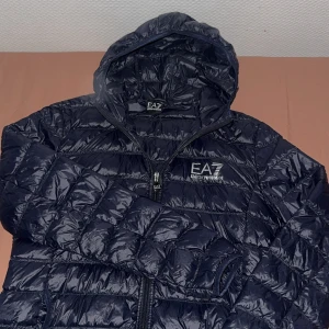 Mörkblå dunjacka EA7 Emporio Armani - Snygg mörkblå dunjacka från EA7 Emporio Armani med glansig finish och tydligt logotryck på bröstet. Jackan har huva, dragkedja framtill och quiltad design. Perfekt för kyliga dagar när du vill hålla stilen.