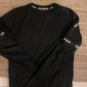 Svart Balmain sweatshirt med logga - Svart sweatshirt från Balmain med broderad logga på bröstet och tryckt Balmain-text längs ärmarna. Tröjan har rund halsringning, ribbade muddar och är tillverkad i mjuk bomull. Perfekt för dig som gillar stilrena designer med tydlig branding.