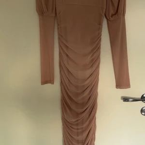 Beige långklänning från Boohoo - Supersnygg beige långklänning från Boohoo med puffiga axlar och långa ärmar. Klänningen har draperad mesh över hela framsidan och en tight passform som framhäver figuren. Perfekt för dig som vill sticka ut med en elegant och trendig look.