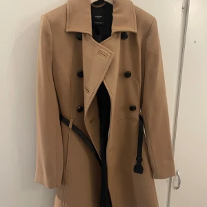 Beige kappa från Mango med bälte XS - Snygg beige kappa från Mango med dubbelknäppning och svart bälte i midjan. Klassisk krage och långa ärmar ger en clean look. Perfekt för dig som vill ha en stilren och tidlös jacka till höst och vinter.