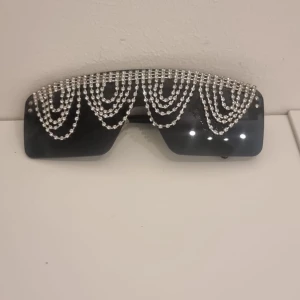 Svarta solglasögon med glittriga kedjor - Unika svarta solglasögon med stora, mörka linser och flera rader av glittrande silverfärgade kedjor över hela ovansidan. Perfekta för dig som vill sticka ut och addera extra bling till din look.