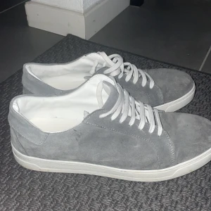Grå mockasneakers från Duca di Morrone - Stilrena sneakers i grå mocka från Duca di Morrone. Skorna har vit sula, vita snören och en klassisk låg profil. Perfekta för dig som gillar en clean och enkel look med en touch av premiumkänsla. Passar till det mesta och är riktigt lätta att matcha.