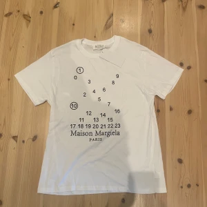 Maison Margiela vit t-shirt med print - Vit t-shirt från Maison Margiela med svart grafiskt print och logga på bröstet. Klassisk rund hals och korta ärmar. Tillverkad i mjuk bomull för en clean och stilren look. Perfekt för dig som gillar minimalistisk design med en twist.