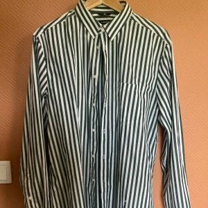 Randig skjorta från J.Lindeberg - Snygg skjorta från J.Lindeberg med breda vertikala ränder i mörkgrönt och vitt. Klassisk krage, knäppning framtill och bröstficka. Långärmad modell i bomull.