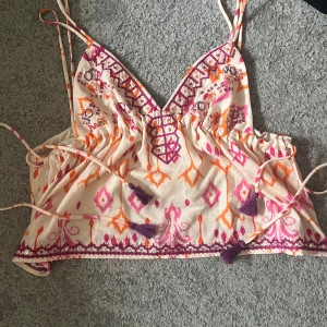 Mönstrad linnetopp med snörning - Superfin linnetopp med tunna axelband och färgglatt mönster i rosa, orange och lila. Toppen har broderade detaljer, snörning med tofsar och v-ringning. Perfekt för sommaren med sitt lätta bomullsmaterial och boho-vibe.