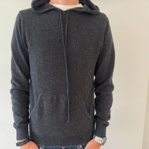 JOHN HENRIC KASHMIR HOODIE - Mysig mörkgrå hoodie i lyxig 100% kashmir från John Henric. Klassisk modell med huva, snörning och känguruficka. Perfekt för dig som gillar en stilren och enkel look med premiumkänsla. Långärmad och riktigt skön mot huden.